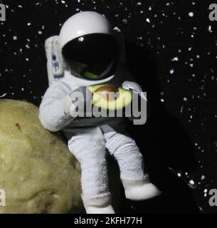 Nahaufnahme einer Figur eines Astronauten, der Avocado auf dem Mond isst, mit einem Sternenhintergrund Stockfoto
