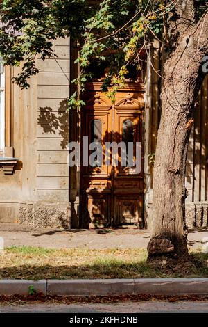 Geschlossene hölzerne Haustür eines Hauses tagsüber. Herbststimmung, verwelkte Blätter auf dem Bürgersteig, der Schatten eines Baumes fällt auf die Tür. Stockfoto