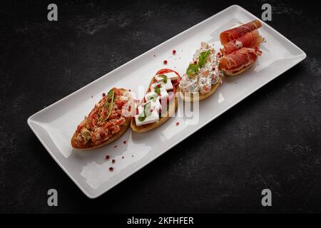Verschiedene Bruschetta mit Jamon, Meeresfrüchten, Gemüse und Käse, auf dunklem Beton Stockfoto