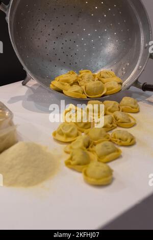 Frisch zubereitete rohe Tortellini, die aus einem Metallsieb austritt, und Mehl auf dem Tisch. Stockfoto