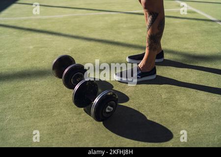 Eine Nahaufnahme der tätowierten Füße eines Mannes neben Hanteln auf grünem Gras in einem Fitnessstudio im Freien Stockfoto