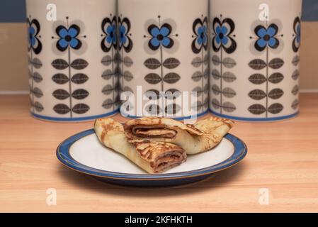 Pfannkuchen auf einem blau-weißen Teller gerollt mit Tee, Zucker und Kaffee Caddy im Hintergrund. Stockfoto