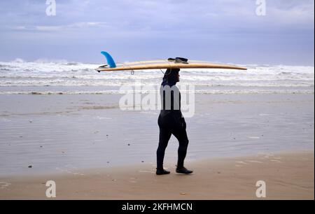 Ein Mann in einem Nassanzug trägt ein Surfbrett auf dem Kopf und läuft am Strand von Saltburn by the Sea, Yorkshire, Großbritannien, entlang. Untersuchungen haben ergeben, dass Reisen in Badeorte der Gesundheit nütze sind. Akademiker von Universitäten befragten Urlauber in Küstenstädten, um nach den Verbindungen zwischen Wohlbefinden und Meer zu fragen. Rund 85 % stimmten ausdrücklich zu oder stimmten darin überein, dass sich das Meer positiv auf Gesundheit und Wohlbefinden auswirkt. Stockfoto