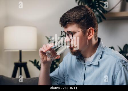 Schöner bärtiger Mann trinkt Wasser aus Glas. Nützliche Gewohnheiten und gesunde Lebensweise Konzept. Der junge Mann in der Brille arbeitet zu Hause auf der Couch sitzend Stockfoto