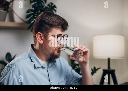 Junger kaukasischer Mann in Gläsern, der zu Hause Wasser trinkt, sitzt auf einer bequemen Couch im Wohnzimmer zu Hause. Schöner bärtiger Kerl trinkt Wasser aus Glas Stockfoto