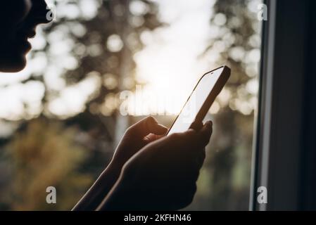 Eine kurze Aufnahme einer jungen Frau, die im Fensterbrett sitzt und das Mobiltelefon hält und die herbstliche Natur während des Sonnenuntergangs genießt. Markt für Smartphone-Anwendungen, Podca Stockfoto