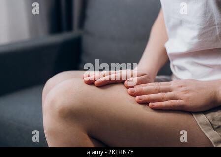 Unkenntlich ungesunde junge Frau mit Schmerzen im Quadrizeps, fühlt sich unwohl, massiert sich auf dem grauen Sofa zu Hause. Nahaufnahme der weiblichen Beine Stockfoto