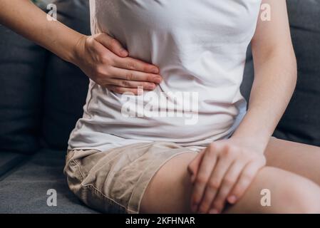 Nahaufnahme eines jungen Weibchens mit Fettleber berührt die rechte Seite mit der Hand, leidet an Bauchschmerzen sitzen auf der grauen Couch. Schmerzen in der rechten Seite, im Blinddarm, Stockfoto