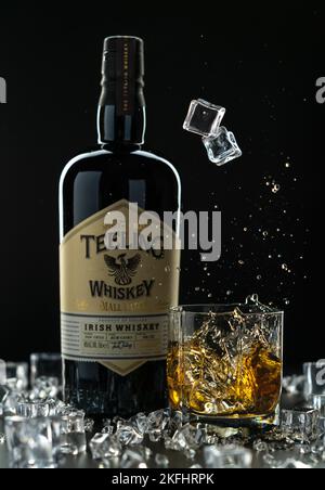 Zwei Eiswürfel mit einem Spritzer fallen in ein Glas mit gegossenem Whiskey auf dem Hintergrund einer Flasche Irish Teeling Whiskey Stockfoto
