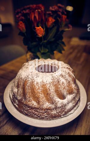 Ein frisch gebackener, hausgemachter, köstlicher gebundener Kuchen, der mit Zuckerpulver bedeckt ist, auf einem Tisch vor Blumen Stockfoto