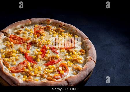 Pizza für Kinder auf Sahnebasis mit Hühnerfilet, Tomaten, Mais und Mozzarella-Käse auf schwarzem Hintergrund. Stockfoto