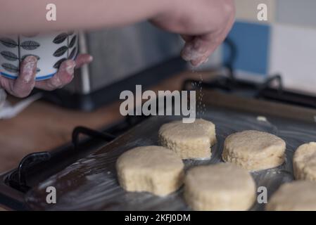 Mann, der Zucker auf ungekochte Scones streut, die er in den Ober tun kann. Auf einem Backblech aus Metall Stockfoto