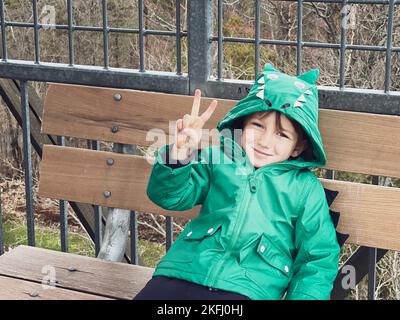 Porträt eines kaukasischen lächelnden Jungen mit grünem Wintermantel und Friedenszeichen, während er am Wochenende auf der Bank im Park sitzt Stockfoto