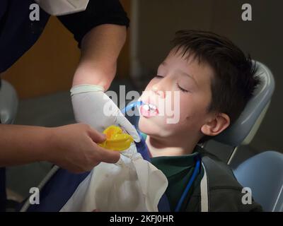 Verkürzte Hände einer Zahnärztin, die einem Jugendlichen Jungen, der im Krankenhaus auf einem Stuhl sitzt, eine medizinische Behandlung gibt Stockfoto