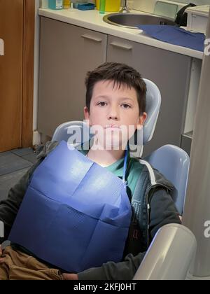 Porträt eines ernsthaften kaukasischen Jungen mit blauem Lätzchen, der in der Zahnklinik auf dem Zahnarztstuhl sitzt Stockfoto