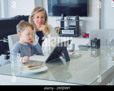 Lächelnde Mutter mit mittlerem Erwachsenen sitzt mit niedlichem Sohn e-Learning durch digitales Tablet auf dem Esstisch zu Hause Stockfoto