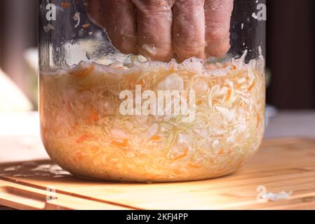 Mann, der hausgemachtes Sauerkraut mit der Hand im Glas heruntertampfte. Fein gehackter Kohl wird aus den Händen in ein Gefäß für weiteren Sauerteig gegossen Stockfoto