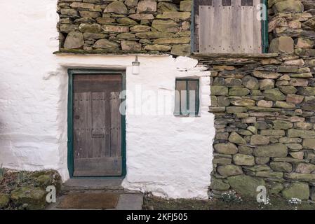 Altes Cottage mit weiß gewaschenen Steinwänden. Alte, rustikale Holztür mit Schneetürenblumen auf dem Boden. Alte rustikale Lampe über der Tür. Stockfoto