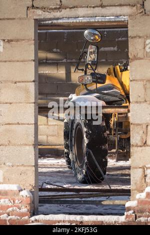 Türöffnung auf der Baustelle mit rostigen Baumaschinen im Gebäude. Gelber Traktor, durch die Tür sichtbar, mit großen Spiegeln. Stockfoto