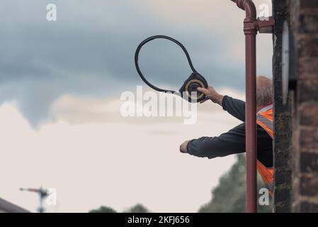 Ein Mitarbeiter der Dampflokomotive, der aus der Signalbox hing und den Token winkte, um ihn dem nächsten Zugfahrer in Goathland, Großbritannien, zu übergeben. Orange, hohe Lichtstärke Stockfoto