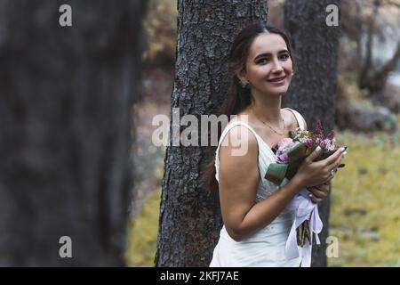 Gerade heiratete sie eine wunderschöne Türkin im Alter von 20s Jahren, die neben dem Waldbaum posierte, lächelnd die Kamera ansah und einen frischen Blumenstrauß in der Hand hielt. Brautkleid. Hochwertige Fotos Stockfoto