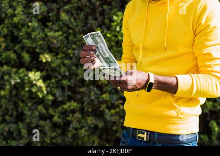 Nahaufnahme der Hände eines afrikanischen Mannes, der in einem gelben Sweatshirt und einem nicht erkennbaren Armband mit Dollarnoten gekleidet ist, während er sie weiterführt Stockfoto