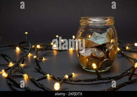 Glasbehälter mit Euro-Münzen und Papierboot auf dunklem Hintergrund mit kleinen LED-Lichtern. Euro-Taschensparkonzept. Stockfoto