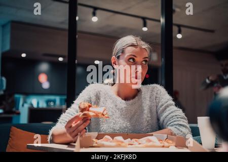 Eine moderne Blondine, die in ihrem Büro Pizza isst, während einer Pause von der Arbeit Stockfoto