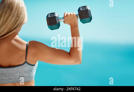 Sie kann das Gewicht auf ihren Schultern tragen. Rückansicht einer nicht erkennbaren jungen Frau, die draußen mit Hanteln trainiert. Stockfoto