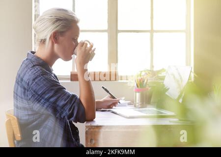 Frische neue Ideen. Eine junge Frau trinkt Kaffee, während sie zu Hause am Computer sitzt. Stockfoto