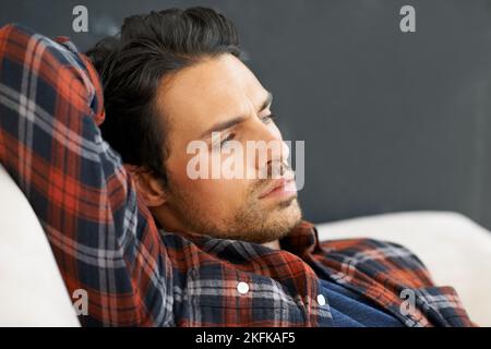 Ich fühle mich brüderlich und launisch. Ein wunderschöner junger Mann, der auf einem Sofa mit einem Arm hinter dem Kopf liegt und mit einem Ausdruck von in die Ferne blickt Stockfoto