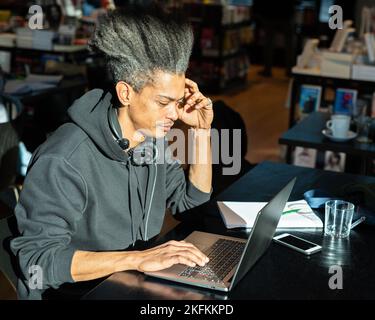 Junger Afro-amerikanischer Mann, der neue Inhalte für seinen persönlichen Blog studiert, junger lateinamerikaner, der an einem Projekt arbeitet, das mit modernen Technologien verbunden ist, an Lebensmut Stockfoto