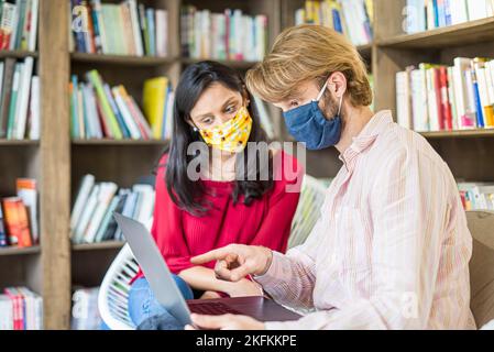Ein junges Paar multirassischer Studenten, die während der Corona Virus-Epidemie einen Laptop in einer Bibliothek benutzten, trug eine Gesichtsmaske zum Schutz, zur Teamarbeit und zur Gesundheit Stockfoto