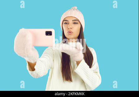 Hübsche Frau im Winterhut und Fäustlinge, die Selfie auf dem Handy machen und Luftkuss schicken Stockfoto