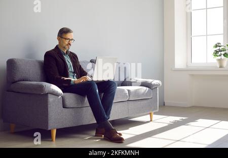 Erfolgreicher reifer Geschäftsmann sitzt auf der Couch und arbeitet an einem modernen Laptop-Computer Stockfoto
