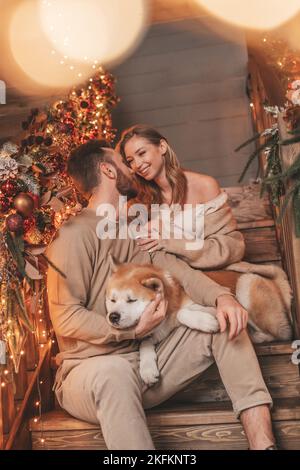 Porträt eines jungen schönen Paares umarmt Indoor Eve 25 Dezember mit akita inu Hund. Menschen in Strick stilvolle Outfit Umarmungen küsst Zärtlichkeit feiert ne Stockfoto
