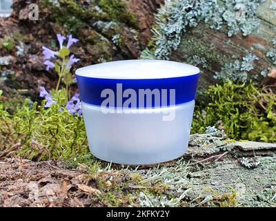 Weißes Kosmetikglas mit blauer Schraubkappe und grünem Waldmoos. Vorderansicht, Nachbau, Nahaufnahme. Kosmetische Flasche Behälter mit Feuchtigkeitscreme für das Branding. Stockfoto