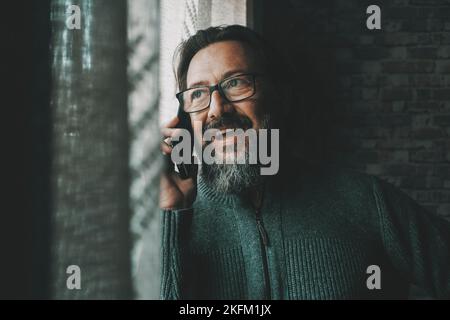 Ein glücklicher junger, reifer Mann, der am Telefon sprach und lächelte und sich zu Hause am Fenster amüsieren musste. Konzept von Menschen und mobiler Zellkommunikation. Männlich Stockfoto
