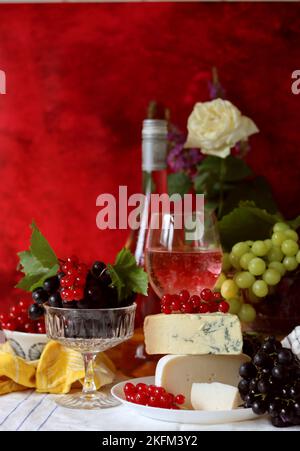Wein, Käse, Obst und Blumen auf einem Tisch. Lebendige Farben frischer Beeren und Trauben. Farbenfrohes Stillleben mit einem Glas rosa Wein und Obst. Stockfoto
