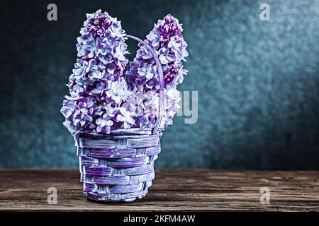 Fliederblüten in einem kleinen Korbkorb auf Vintage-Holz und schwarzem Hintergrund Stockfoto