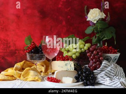 Wein, Käse, Obst und Blumen auf einem Tisch. Lebendige Farben frischer Beeren und Trauben. Farbenfrohes Stillleben mit einem Glas rosa Wein und Obst. Stockfoto