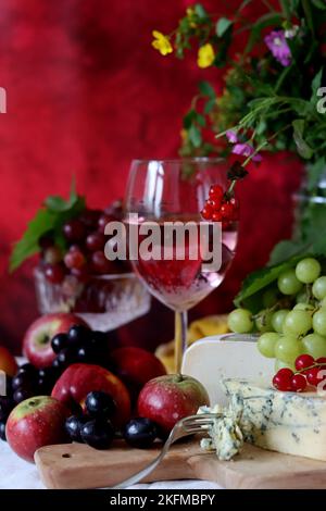 Wein, Käse, Obst und Blumen auf einem Tisch. Lebendige Farben frischer Beeren und Trauben. Farbenfrohes Stillleben mit einem Glas rosa Wein und Obst. Stockfoto