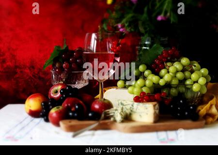 Wein, Käse, Obst und Blumen auf einem Tisch. Lebendige Farben frischer Beeren und Trauben. Farbenfrohes Stillleben mit einem Glas rosa Wein und Obst. Stockfoto