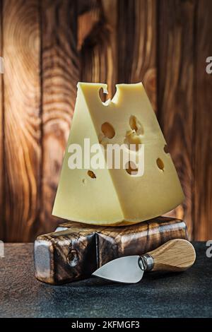Vertikale Ansicht großes Stück Milch maasdam Käse auf kleinen Schneidebrett und Messer alten Holzhintergrund Stockfoto