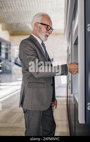Seitenansicht eines älteren männlichen professionellen Mannes, der einen Geldautomaten benutzt. Leitender Geschäftsmann mit Brille, während er Bargeld vom Geldautomaten abzieht. Er trägt s Stockfoto