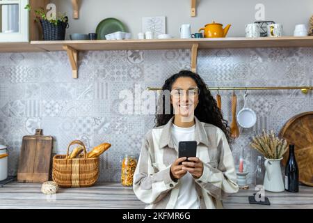 Eine schöne junge Lateinamerikanerin hält ein Telefon in den Händen. Zu Hause in der Küche stehend, lächelnd auf die Kamera schauend. Stockfoto