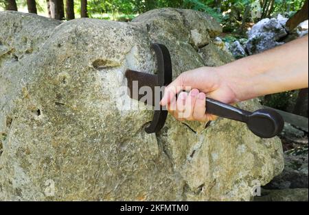 Die Hand des jungen Mannes, der gewaltsam versucht, das Schwert Excalibur zu extrahieren, steckt wie König Arthur im Felsen fest Stockfoto