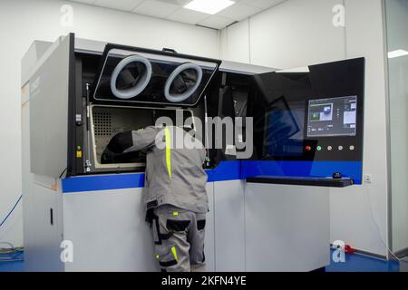 Ein Arbeiter blickt in ein offenes Fenster eines großen industriellen 3D-Druckers für Metallpulver. Der Arbeitsprozess. Industrial Powder 3D Drucker für Metall und Arbeiter Mann. Laser-Sintermaschine für Metall. Stockfoto