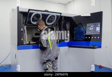 Ein Arbeiter blickt in ein offenes Fenster eines großen industriellen 3D-Druckers für Metallpulver. Der Arbeitsprozess. Industrial Powder 3D Drucker für Metall und Arbeiter Mann. Laser-Sintermaschine für Metall. Stockfoto