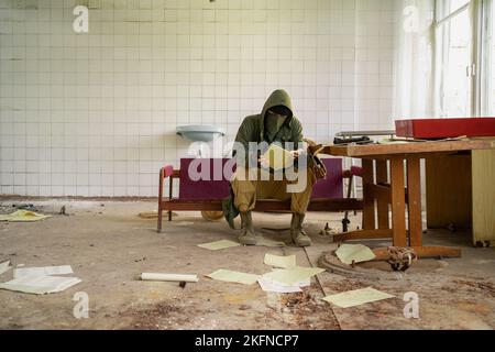 Mann in Bandana-Maske, Kapuze und Camo-Hose sitzt in verlassenen Gebäude und liest Dokumente. Cyberpunk postapocalypse Fantasy Horrorszene, oder ai Stockfoto